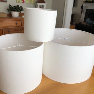 May include: Three white fabric lampshades of different sizes. The lampshades are cylindrical and have a textured surface.