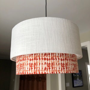 May include: A white drum lampshade with a red and orange patterned inner shade. The lampshade is hanging from a black cord.