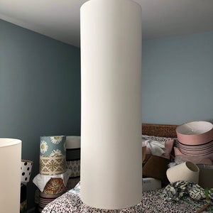May include: A white cylindrical lampshade hangs from the ceiling in a bedroom. The lampshade is large and appears to be made of fabric. The room is decorated with a variety of patterned lampshades and a bed with a floral patterned duvet.