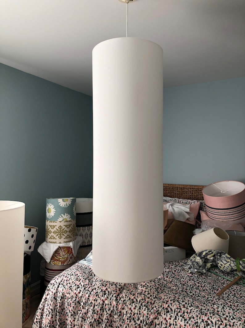 May include: A white cylindrical lampshade hangs from the ceiling in a bedroom. The lampshade is large and appears to be made of fabric. The room is decorated with a variety of patterned lampshades and a bed with a floral patterned duvet.