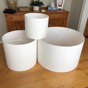 May include: Three white cylindrical lampshades of different sizes are arranged on a wooden surface. The shades are made of a smooth, opaque fabric. The largest shade is at the base, with the smallest one stacked on top. The shades are of varying heights.