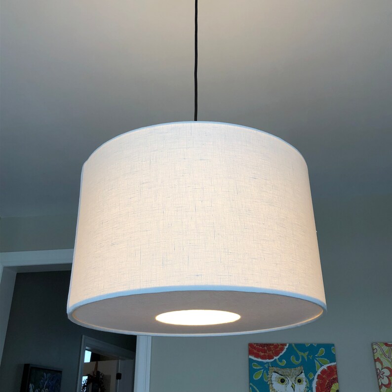 May include: A white drum-shaped lampshade with a white fabric covering and a white trim around the bottom edge. The lampshade is hanging from a black cord.