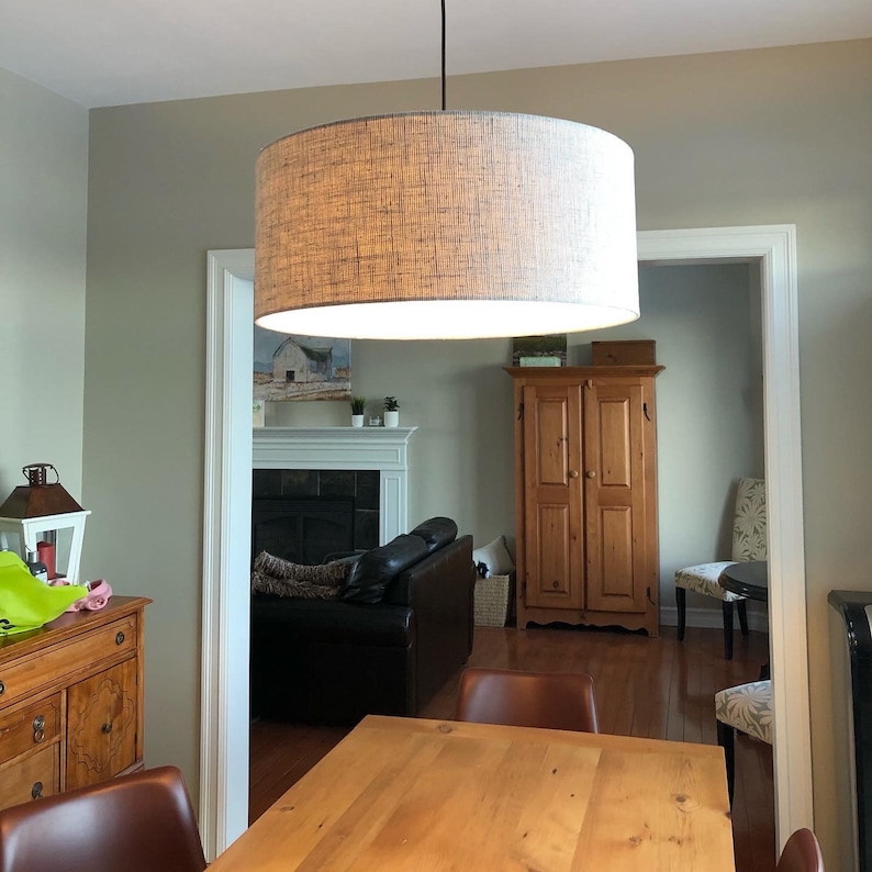 May include: A brown woven drum shade pendant light fixture hangs from the ceiling over a wooden dining table. The light fixture is in a dining room with a fireplace and a wooden cabinet in the background.