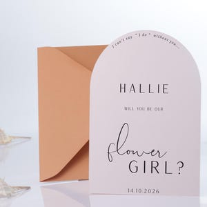 May include: A pink card with rounded edges and a white arch design. The card says "I can't say 'I do' without you..." and "HALLIE, will you be our flower girl?" with the date "14.10.2026".