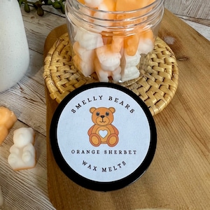 May include: A glass jar filled with orange and white wax melts shaped like bears. The jar has a black lid with a white label that says "Smelly Bears Orange Sherbet Wax Melts". The label also features a cartoon bear with a heart on its chest.