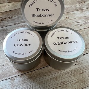 May include: Three silver candle tins with white labels. The labels read "HIGHLY THRIFTED CO." and feature different Texas-themed scents: "Texas Bluebonnet", "Texas Cowboy", and "Texas Wildflowers". Each tin contains 3 ounces of natural soy wax.
