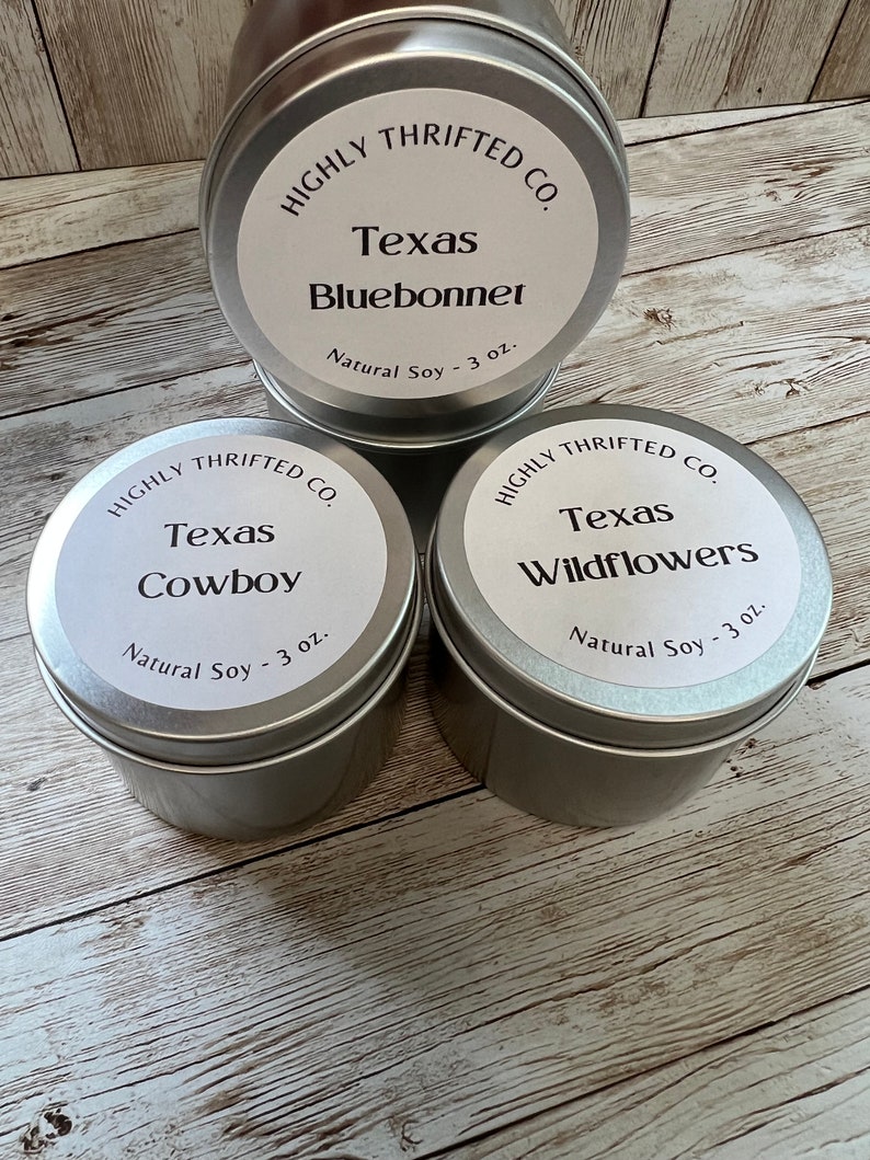 May include: Three silver candle tins with white labels. The labels read "HIGHLY THRIFTED CO." and feature different Texas-themed scents: "Texas Bluebonnet", "Texas Cowboy", and "Texas Wildflowers". Each tin contains 3 ounces of natural soy wax.