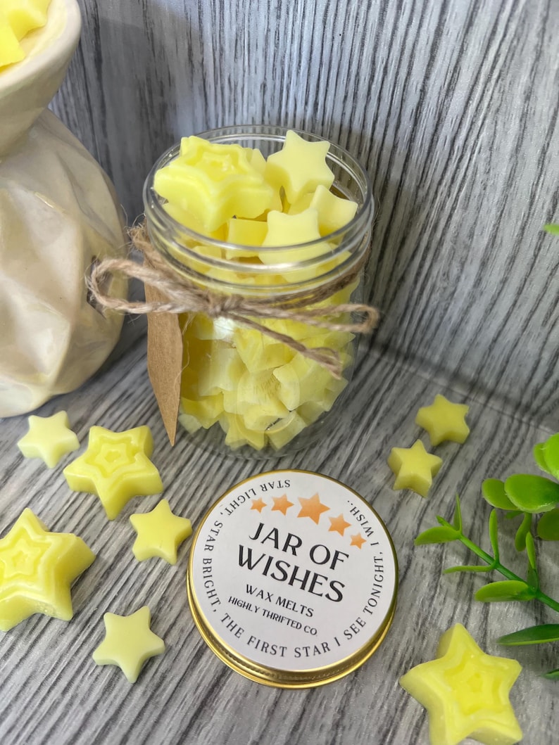 May include: A glass jar filled with yellow star-shaped wax melts. The jar is tied with twine and a brown paper tag. The jar is sitting on a wooden surface with a gold lid that says "Jar of Wishes Wax Melts Highly Thrifted Co The First Star I See Tonight."