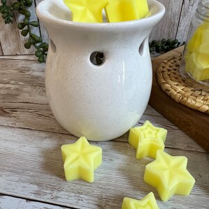 May include: White ceramic wax warmer with a speckled finish and five yellow star-shaped wax melts. The warmer has a round base and a wide opening at the top. The wax melts are arranged in the warmer and on the wooden surface.