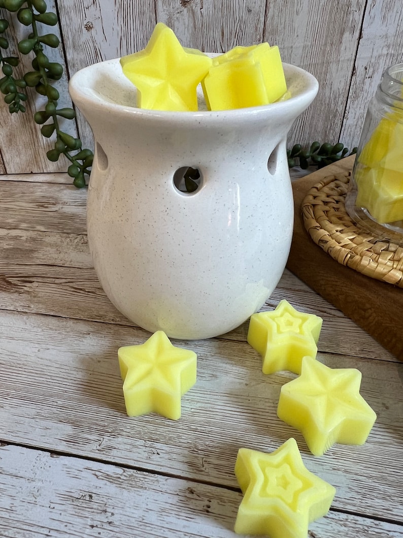 May include: White ceramic wax warmer with a speckled finish and five yellow star-shaped wax melts. The warmer has a round base and a wide opening at the top. The wax melts are arranged in the warmer and on the wooden surface.