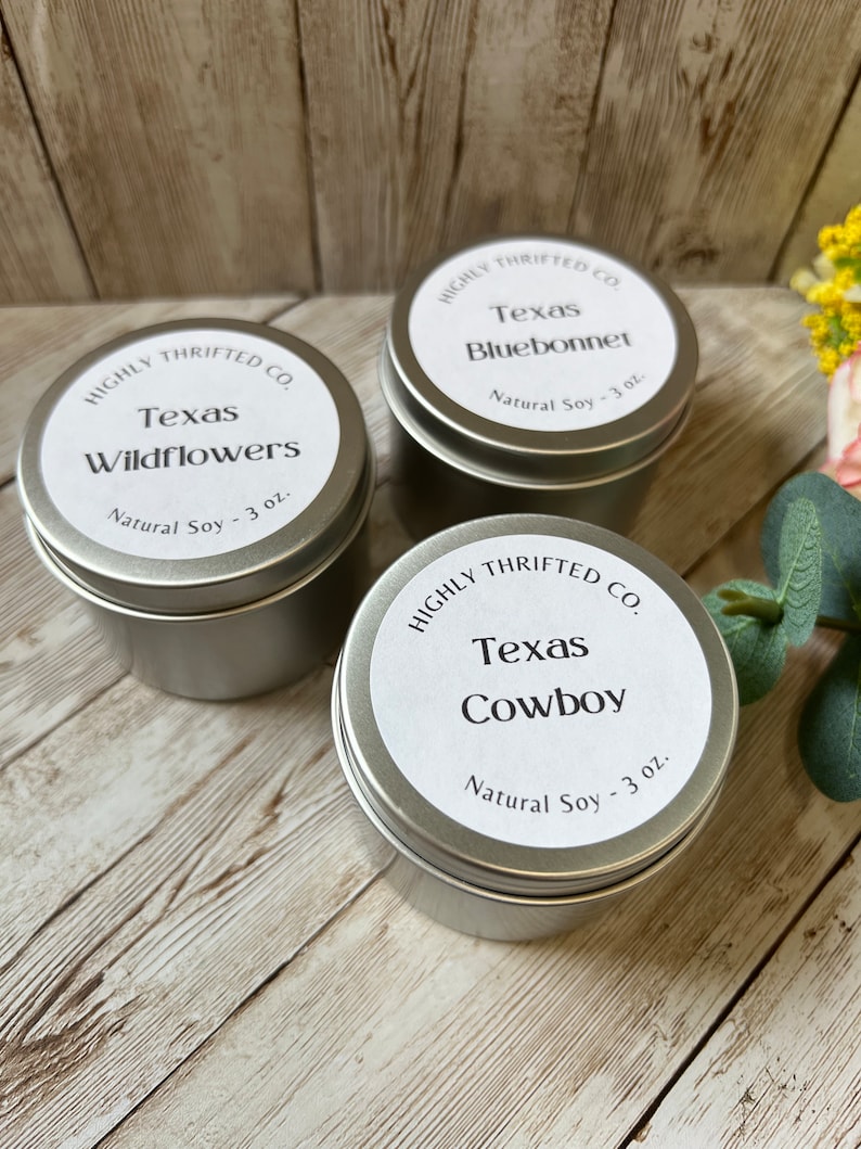May include: Three silver candle tins with white labels. Each label has the text "HIGHLY THRIFTED CO." and a different Texas-themed scent: "Texas Wildflowers", "Texas Bluebonnet", and "Texas Cowboy". Each tin contains 3 ounces of natural soy wax.