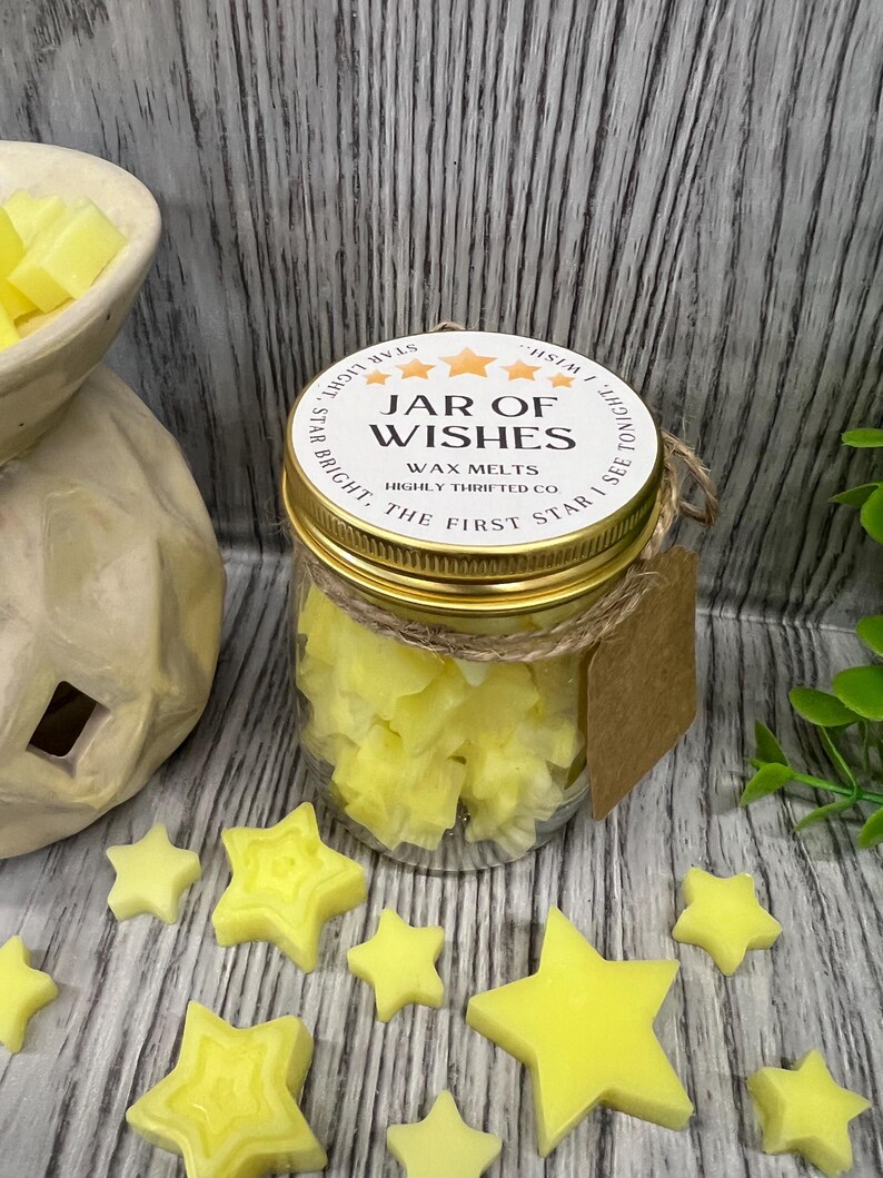 May include: A glass jar filled with yellow star-shaped wax melts. The jar has a gold lid with the text "JAR OF WISHES WAX MELTS HIGHLY THRIFTED CO. STAR BRIGHT, THE FIRST STAR I SEE TONIGHT."