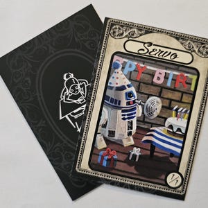 May include: Two cards are shown. One card has a birthday scene with a robot wearing a party hat, a cake, and the words "Happy Birthday." The other card is black with a white illustration.