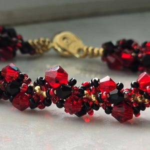 May include: A bracelet made of red and black beads with gold-coloured accents. The beads are faceted and clustered together. The bracelet is fastened with a gold-toned clasp. The design creates a textured appearance.