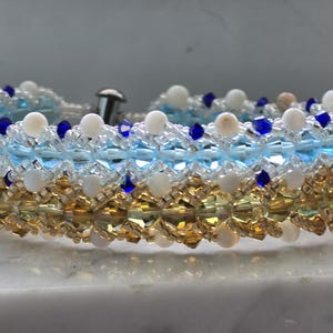 May include: A beaded bracelet with white, blue, and gold beads. The bracelet has a clasp and is designed with multiple layers of beads. The beads are arranged in a decorative pattern.