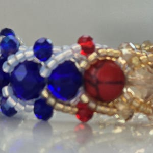 May include: A beaded bracelet featuring blue, white, gold, and red beads. The bracelet is woven in a pattern and has a red heart-shaped bead at the centre.