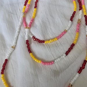 May include: A multi-strand beaded waist chain in various colours. The beads are white, yellow, pink, and red. The waist chain is displayed on a white fabric background.