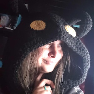 May include: A black crochet hood with two rounded ears and two tan-colored circular accents. The hood is worn by a person. The hood is made of a thick, textured yarn and has a cozy appearance.