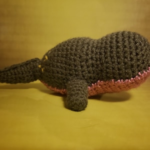 May include: A crocheted plush toy shaped like a whale. The whale is dark green with a pink underside and yellow accents.