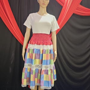 May include: A mannequin wearing a white short-sleeved t-shirt and a colorful patchwork skirt. The skirt has a red waistband and is trimmed with white lace. A white head wrap completes the outfit. The background features red fabric draped over a black backdrop.