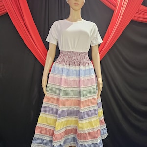 May include: A mannequin wearing a white t-shirt and a colorful patchwork skirt. The skirt features alternating bands of gingham fabric in various colors, including red, blue, yellow, and green, with white lace trim. A white headscarf completes the outfit.