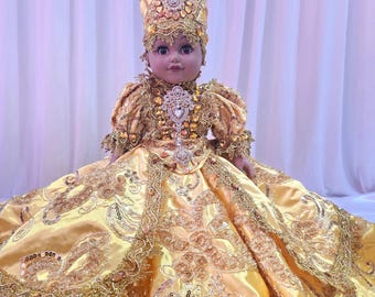 May include: A doll dressed in a golden yellow gown and matching ornate headdress. The gown features intricate gold embroidery and embellishments. The doll's face is detailed with painted features, and it is posed against a soft, white background.