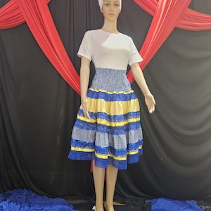 May include: A mannequin wearing a white short-sleeved top and a blue, yellow, and white skirt. The skirt has a checkered pattern on the top, with yellow and blue stripes below. A white head wrap and a flower complete the outfit. The background features red and black fabric.