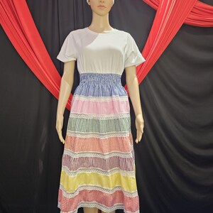 May include: A mannequin displays a colorful, tiered dress. The top is white, transitioning to a blue gingham waist. The skirt features alternating layers of pink, green, orange, yellow, and red gingham, each separated by white lace trim. A white head wrap completes the outfit.
