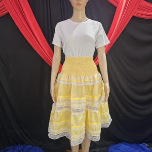 May include: A white short-sleeved top paired with a yellow skirt. The skirt features a smocked waistband, yellow fabric, white lace trim, and yellow ribbon accents. A white head wrap with a flower completes the outfit.