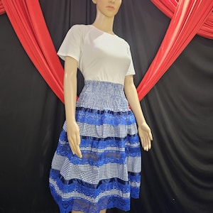 May include: A mannequin wearing a white short-sleeved top and a blue and white checkered skirt. The skirt has blue lace trim and blue ribbon accents. A white headband with a flower is on the mannequin's head.