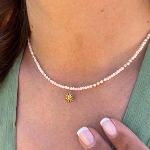 Può includere: Una delicata collana con una fila di piccole perline di colore chiaro. Un ciondolo a forma di sole dorato con accenti gialli pende al centro. La collana è indossata su un top verde.