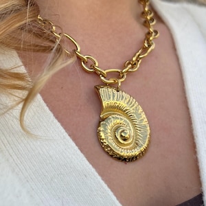 Peut inclure: Un collier doré avec un grand pendentif détaillé en forme de coquille de nautile. Le pendentif est suspendu à une grosse chaîne. Le bijou est porté sur un pull blanc.