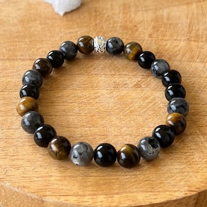 Peut inclure: Un bracelet en perles avec des pierres noires, brunes et grises. Le bracelet a une perle argentée au centre.