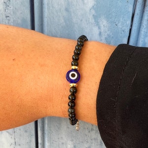 Peut inclure: Bracelet en perles noires avec un charme œil bleu et blanc. Le bracelet est au poignet d'une personne.