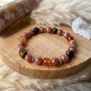 Puede incluir: Un brazalete hecho con cuentas de cornalina, ojo de tigre y piedra de sol. Las cuentas están separadas por separadores de color dorado. El brazalete está sobre una superficie de madera.
