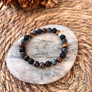 Peut inclure: Un bracelet composé de perles de lave noire, d'œil de tigre et de pierres précieuses grises. Le bracelet est orné d'une perle argentée avec un motif décoratif.