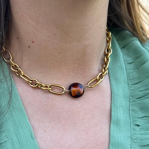 Peut inclure: Collier chaîne doré avec un grand pendentif rond en forme d'écaille de tortue. Le collier est porté sur un haut vert. Les maillons de la chaîne sont ovales et épais. Le pendentif est de couleur marron foncé et ambre.