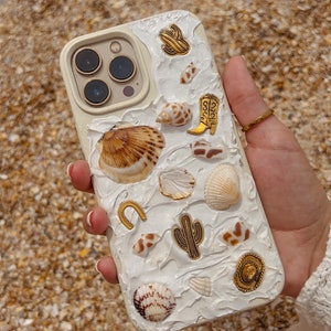 May include: White iPhone case decorated with seashells, cacti, and gold accents. The case has a textured, 3D effect.