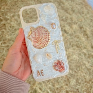 May include: White iPhone case with a seashell design. The case has a variety of seashells, including a large pink and white seashell, a small white seashell, and a pearl. The case also has gold accents, including a gold bow and the letters "AE".