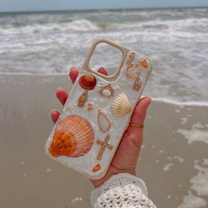 May include: A white phone case decorated with seashells, gold accents, and a small cross. The case is being held in a hand with a white crocheted sleeve.