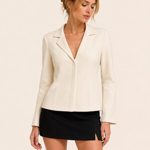May include: Cream-colored blazer with a notched lapel and long sleeves. The blazer is paired with a black mini skirt. The skirt has a side slit. The outfit is suitable for business or casual wear.