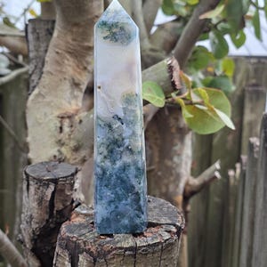 May include: A green, white, and blue agate crystal point, also known as a wand, is sitting on a tree stump. The crystal is polished and has a pointed tip.