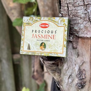 May include: A box of "PRECIOUS JASMINE" incense cones. The box is light green and gold with the brand "HEM" in a red circle. The box is resting on a tree trunk.