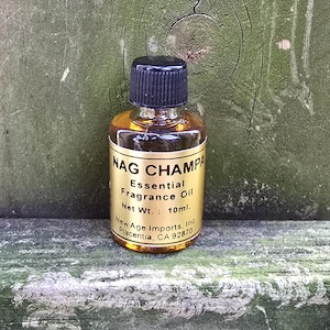 May include: A small glass bottle of Nag Champa essential fragrance oil. The bottle has a black cap and a gold label with black text. The label reads "Nag Champa Essential Fragrance Oil Net Wt.: 10ml."