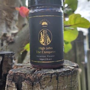 Puede incluir: Un frasco negro con letras doradas que dice "High John The Conqueror Incense Power 50gm (1.76 oz.)" y una imagen dorada de un rey. El frasco está sentado en un tocón de árbol.