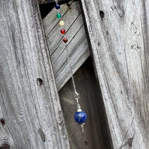 Op de afbeelding: Een pendel met een zilveren ketting en een blauwe lapis lazuli bol aan de onderkant. De ketting is bevestigd aan een reeks kleurrijke edelsteen kralen. De achtergrond bestaat uit verweerde houten planken.
