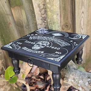 Könnte beinhalten: Schwarzer Holztisch mit Ouija-Brett und einem Totenkopf- und Handdesign. Der Tisch hat vier Beine und eine kleine Schublade mit einem Ringzug.