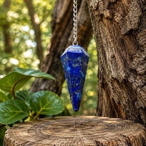 May include: A faceted, deep blue lapis lazuli pendulum with natural gold-coloured pyrite inclusions, suspended from a silver-toned metal chain. The gemstone is shaped into a pointed, multi-sided geometric form, showcasing a smooth, polished surface.