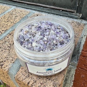 May include: A clear plastic jar filled with amethyst sand. The label reads "Vrinda's Amethyst Sand 200g / 7.05oz".
