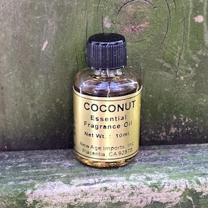 Puede incluir: Una pequeña botella dorada con una tapa negra etiquetada "COCONUT Essential Fragrance Oil Net Wt.: 10ml. New Age Imports, Inc. Placentia, CA 92870"